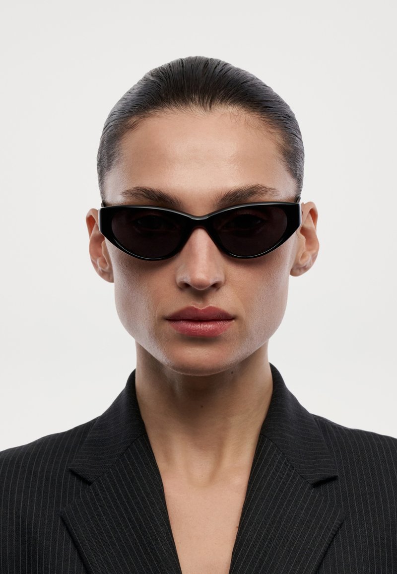 Femme aux cheveux tirés en arrière portant des lunettes de soleil noires forme œil de chat et un blazer à fines rayures foncé sur un fond clair uni.