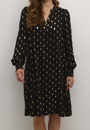 Žena nosící černé dlouhé šaty se zlatými oválnými vzory, výstřihem do V a nařasenými manžetami, stojící u jednobarevného pozadí.