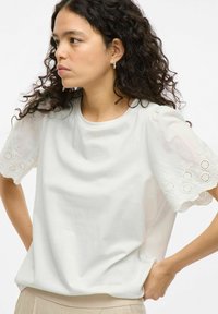 Camiseta de algodón blanca con mangas cortas abullonadas que presenta recortes florales bordados. Textura suave y corte relajado.