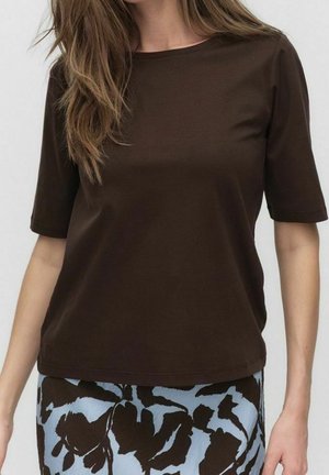 Nainen pukeutunut tummanruskeaan lyhythihaiseen puseroon ja siniseen hameeseen, jossa on suuria tummanruskeita kukkakuoseja.