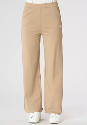 Pantaloni beige a gamba larga con orli spaccati indossati con sneakers bianche e top bianco infilato, mostrati dalla vita ai piedi.