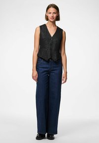 Kvinna står, klädd i en ärmlös svart texturerad väst, mörkblå vida jeans och svarta skor mot en vit bakgrund.