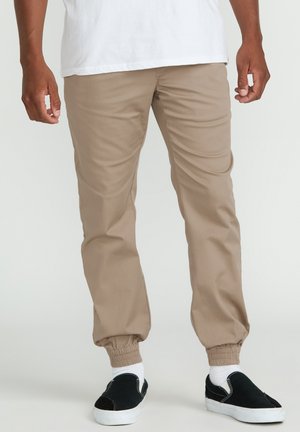 Homme portant un pantalon de jogging beige, des chaussettes blanches, des baskets noires sans lacets et un t-shirt blanc, debout sur un fond clair uni.