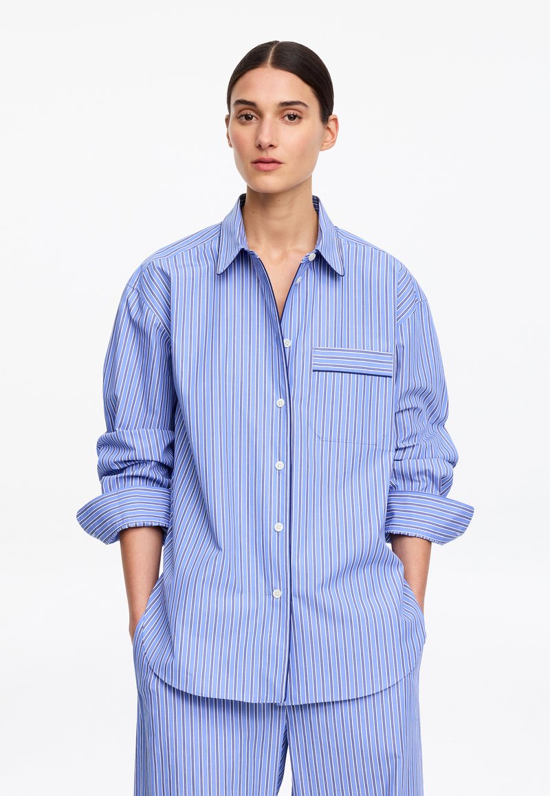 Femme portant une chemise ample bleu clair et un pantalon assorti avec des rayures blanches et rouges, debout les mains dans les poches.