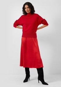 Czerwony dziergany sweter z wysokim kołnierzem i opuszczonymi ramionami, zestawiony z długą, zwiewną czerwoną spódnicą. Modelka nosi czarne botki na obcasie.