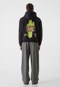 Črna kapuca s zelenim grafičnim motivom kaktusa in napisom "WESTERN MOTEL" na hrbtni strani. Obleko dopolnjujejo ohlapne sive hlače.