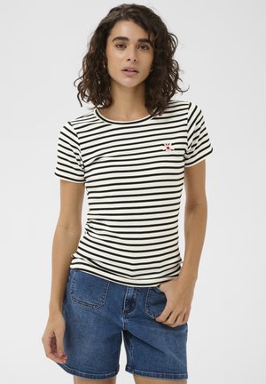 Sieviete ar cirtainiem matiem, tērpusies melnbaltā svītrotā īsroku krekliņā ar mazu ziedu rakstu un zilās džinsa šortās, stāvot pret balto fonu.