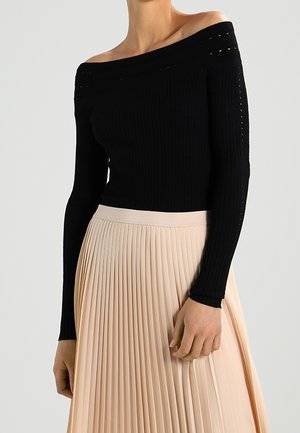 Femme portant un pull côtelé noir épaules dénudées et une jupe plissée beige taille haute, montrée du cou jusqu'au milieu des cuisses.