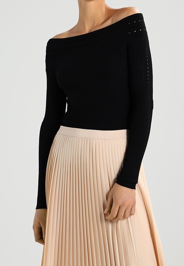 Femme portant un pull côtelé noir épaules dénudées et une jupe plissée beige taille haute, montrée du cou jusqu'au milieu des cuisses.