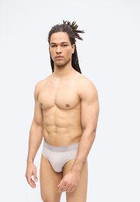 Izmos férfi rasztahajjal, világosszürke Calvin Klein alsóneműt viselve, egyszínű fehér háttér előtt áll, szemben a kamerával.