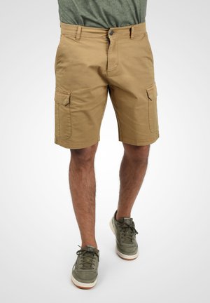 Shorts cargo beiges avec deux poches latérales, en tissu en mélange de coton. Dotés d'une fermeture à bouton et d'un ourlet droit. Portés avec des baskets vertes.