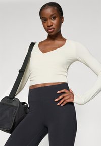 Licht crèmekleurig crop top met lange mouwen en een v-hals, gecombineerd met zwarte high-waisted leggings. Model houdt een zwarte sporttas met een schouderband vast.