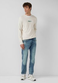 Camicetta a maniche lunghe color crema con logo verde, abbinata a jeans azzurri chiari e sneakers grigie con accenti e lacci bianchi.