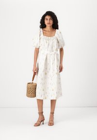 Vestido midi estampado floral em branco, com mangas bufantes e decote franzido. Inclui um cinto atado. Bolsa de cesta trançada complementa o visual.