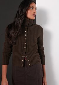 Cardigan marrone a coste con colletto alto, caratterizzato da bottoni sul davanti e maniche lunghe. Abbinato a una gonna marrone scuro.