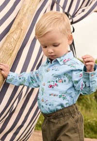 Camicia azzurro chiaro con motivi di veicoli, bottoni e maniche lunghe; abbinata a pantaloni verde oliva con vita elastica. Lo sfondo presenta un tessuto a righe.
