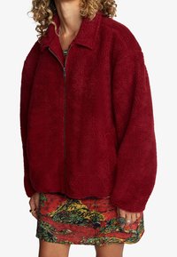 Maroon fleecejacka med dragkedja fram, spetsig krage och oversized passform. Texturerad yta och sänkta axlar. Kombinerad med en färgglad mönstrad kjol.