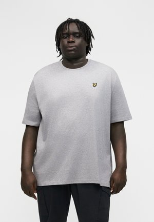 Mladý muž s tmavou pokožkou a dredmi, oblečený v svetlošedom tričku s malým žltým motýlikom, stojaci pred jednoduchým pozadím.