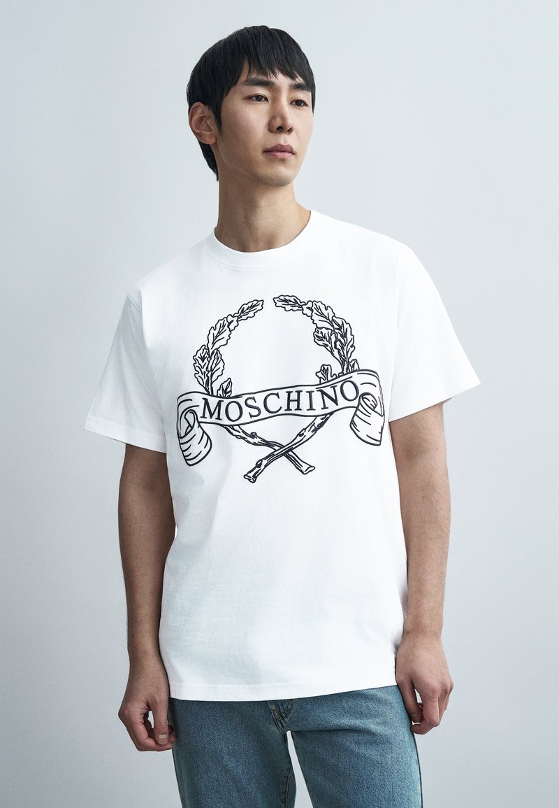 Λευκό βαμβακερό t-shirt με μαύρο γραφικό τύπωμα ενός στεφάνου δάφνης και διασταυρωμένων αντικειμένων με τη λέξη "MOSCHINO" σε κορδέλα στο κέντρο.