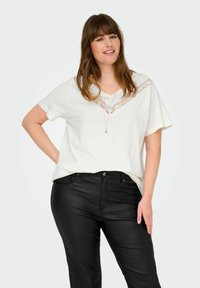 Bílý krátký rukáv top s výstřihem do V a krajkovými detaily, v kombinaci s černými přiléhavými kalhotami, které ukazují elegantní a hladkou texturu.