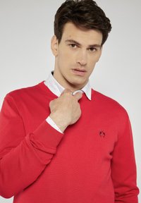 Czerwony sweter wykonany z dzianiny, z okrągłym dekoltem i ściągaczami na rękawach. Pod spodem noszona jest biała koszula z delikatnym wzorem.