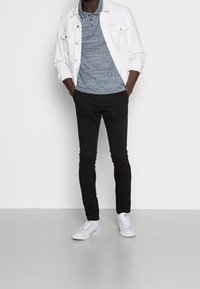 Veste en denim blanche sur une chemise polo bleue à motifs, associée à un pantalon noir slim et des baskets blanches. Design simple, lignes épurées.