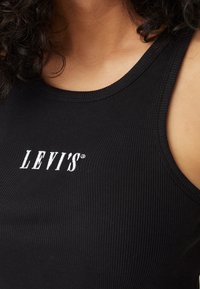 Lähellä otettu kuva henkilöstä, joka pukeutuu mustaan ribbited tanktoppiin, jossa on valkoinen "LEVI'S"-logo brodeerattu rinnassa.