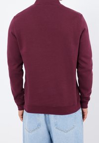 Bordó mikina s dlouhými rukávy, žebrovanými manžetami a lemem, vyrobená z hladké bavlněné směsi. Pohled zezadu odhaluje minimalistický design.