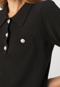 Czarny dzianinowy polo z kołnierzykiem, z pięcioma perłowymi guzikami oraz kieszenią z podobnym ozdobnym guzikiem. Gładka tekstura.