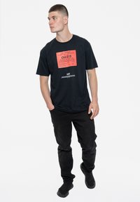 Black cotton t-shirt featuring a prominent red graphic reading "Oasis" and concert details. Paired with black pants and black footwear.
