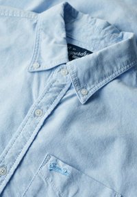 Jasnoniebieska koszula na guziki wykonana z tkaniny o miękkiej fakturze. Posiada spiczasty kołnierzyk, dwa guziki z przodu oraz małą kieszonkę z logo.