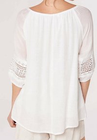 Blouse blanche à coupe décontractée, dotée de manches trois-quarts et d'un détail en dentelle délicate aux poignets. Tissu léger et texturé.