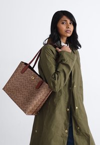 Brown patterned tote bag with red accents, gold hardware, and sturdy handles. Model wears a green coat, holding the bag on her shoulder.