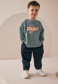 Light blue sweatshirt with airplane graphic, striped cuffs; navy loose-fitting pants; white sneakers; neutral background.
