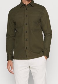 Dunkelolivgrünes Hemd zum Knöpfen mit zwei Brusttaschen, schwarzen Knöpfen und klassischem Kragen; gefertigt aus einem glatten, strukturierten Material.