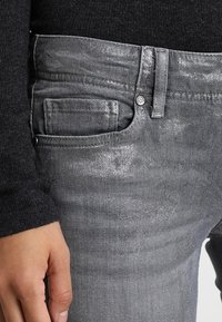 Los jeans de mezclilla gris presentan un acabado elegante y ligeramente brillante, con un bolsillo frontal y un cierre clásico de botón. La tela tiene una apariencia suave y texturizada.