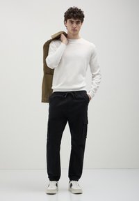 Junger Mann mit lockigem Haar, der einen weißen Pullover, schwarze Cargo-Hosen und weiße Turnschuhe trägt und eine olivfarbene Jacke über seiner Schulter hält.