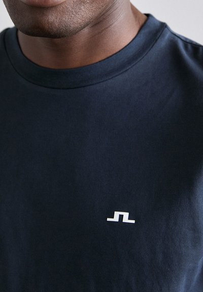 Camiseta de algodón de cuello redondo en color azul marino, con un pequeño logo abstracto blanco en la parte izquierda del pecho, textura suave y corte estándar.