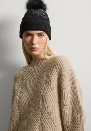 Black knit beanie with a pom-pom, featuring floral embroidery. Cream-colored oversized sweater with textured diagonal patterns and ribbed neckline.