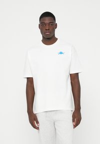 Junger Mann in einem schlichten weißen T-Shirt mit kurzen Ärmeln und kleinem blauen Logo auf der Brust sowie hellgrauen Sweatpants vor weißem Hintergrund.