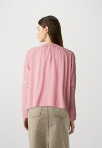 Blusa a maniche lunghe di colore rosa chiaro con scollatura arricciata e dettagli a pettine nella parte posteriore, realizzata in un tessuto morbido, abbinata a una gonna beige.