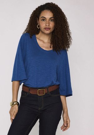 Vrouw met krullend haar die een blauw top met driekwart mouwen draagt, donkere spijkerbroek, een bruine riem, gouden oorringen en een gouden armband.