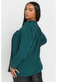 Blusa teal con maniche lunghe, colletto e dettagli con borchie sui polsini. Presenta una vestibilità comoda e una piega sul retro per un tocco di stile in più.