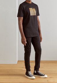 Man wearing black Morato T-shirt with gold stripes, black fitted pants, and black high-top Converse sneakers standing on wooden floor.