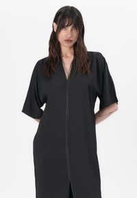 Black, short-sleeve dress with a front zipper closure. Made of a smooth, lightweight fabric with a relaxed fit and side slits.