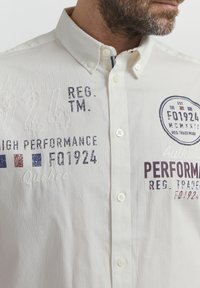 Camisa blanca de botones con texto y logotipos bordados en azul, rojo y gris. Textura suave con un diseño de cuello y cierre de botón.