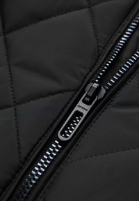 Sort quiltet stof med et nærbillede af en lynlås med en sølvmetal trækstang og tænder. Teksturen ser glat og holdbar ud.