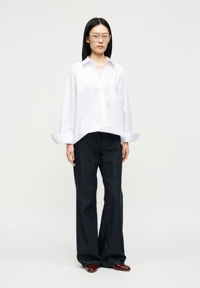 Femme portant une chemise blanche à boutons oversize, un pantalon large sombre, des chaussures bordeaux et des lunettes, debout devant un fond clair uni.