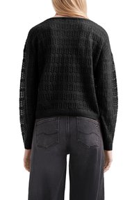 Vrouw in een zwart gebreid sweater met lange mouwen en bloemenpatronen, gedragen met vervaagde zwarte jeans, weergegeven vanuit de achterkant tegen een witte achtergrond.