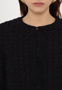 Femme portant un haut noir texturé avec un col rond et des liens à l'avant, avec une peau claire visible et des cheveux bruns lâches derrière les oreilles.